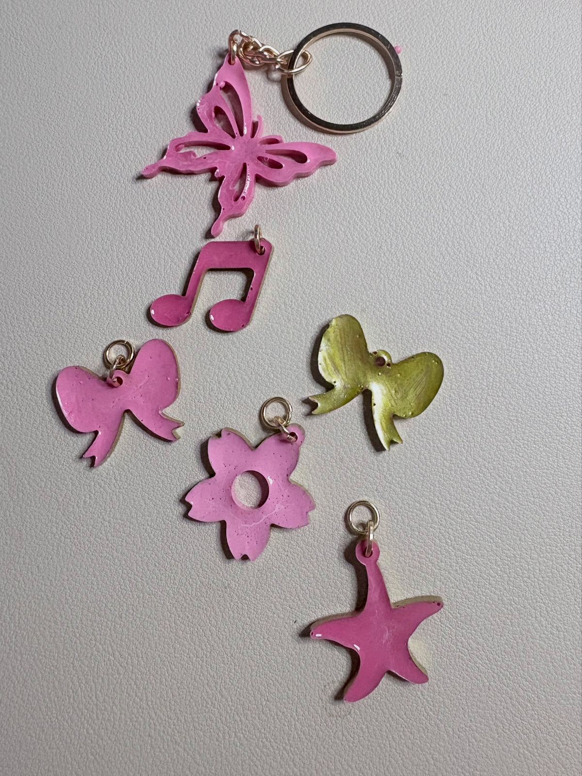A set of six enamel keychain charms in pink and gold, including a butterfly, music note, bow, flower, starfish, and a small bird, arranged on a light gray textured background.