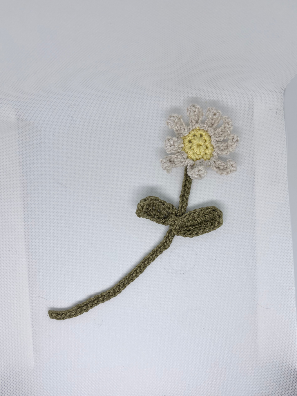 A handmade crocheted flower with a white and yellow face, an olive green stem, and a matching bow, displayed on a white textured background.