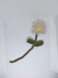 A handmade crocheted flower with a white and yellow face, an olive green stem, and a matching bow, displayed on a white textured background.