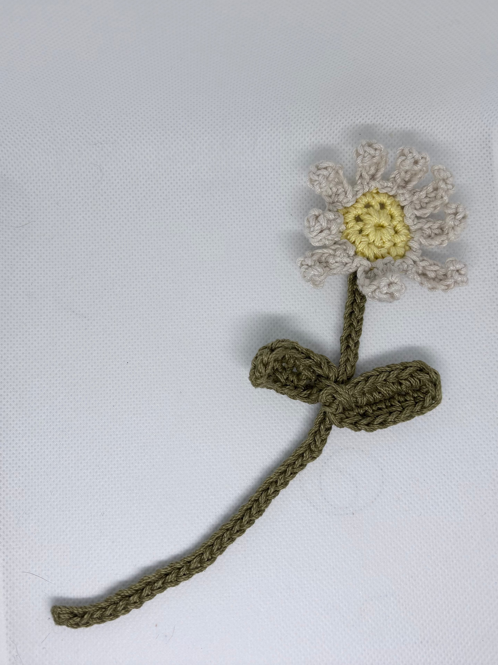 A small crocheted flower accessory with an olive green stem and a daisy-like head in off-white with a yellow center, placed on a white textured surface.
