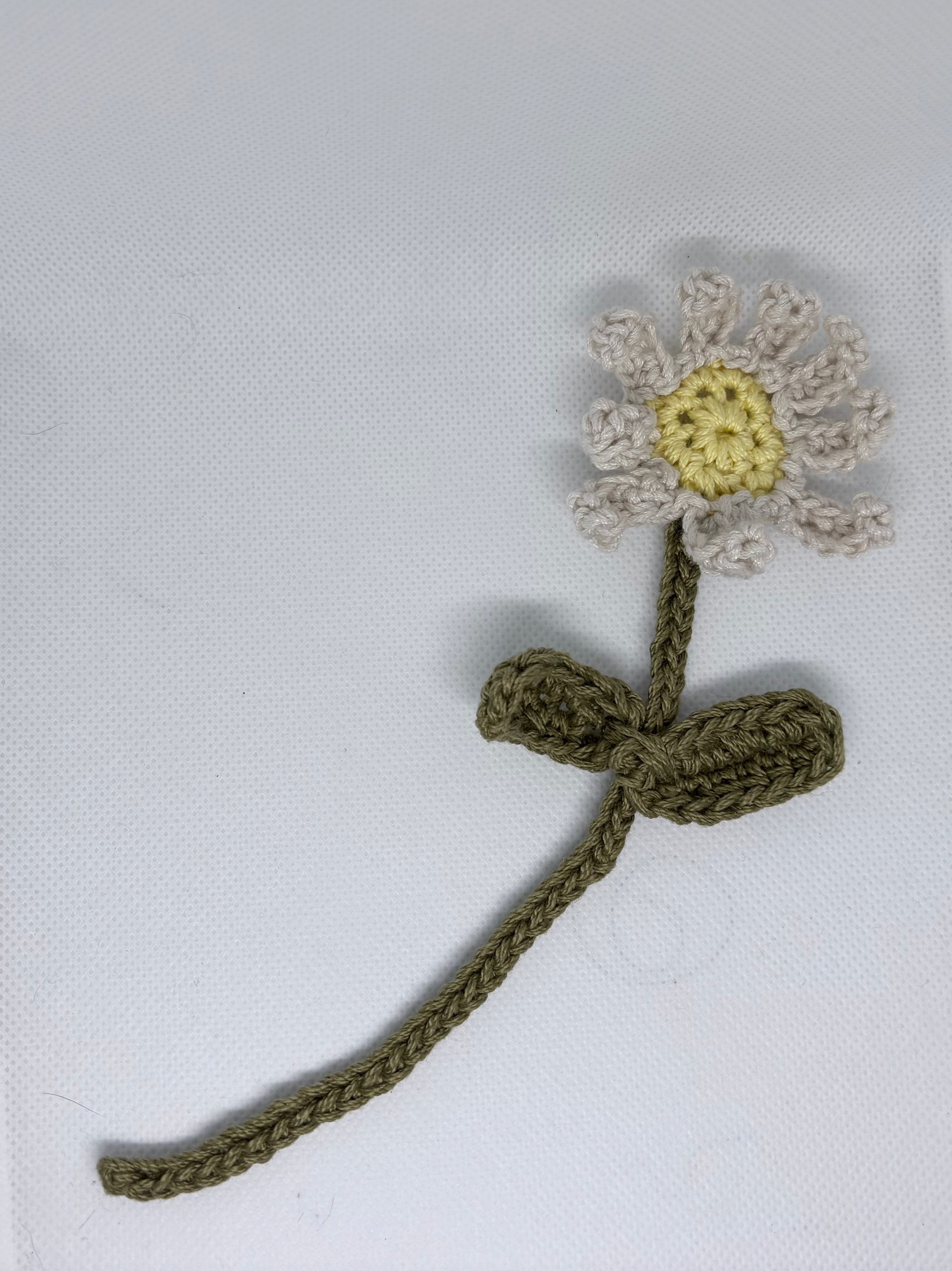 A small crocheted flower accessory with an olive green stem and a daisy-like head in off-white with a yellow center, placed on a white textured surface.
