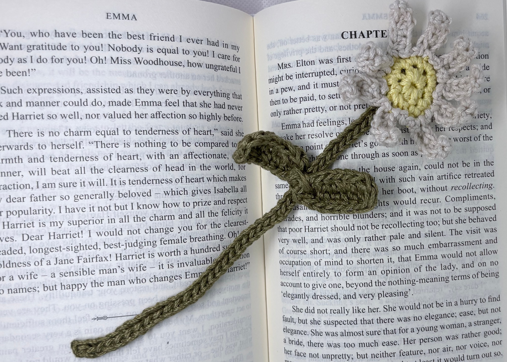 A handmade crocheted bookmark shaped like a flower with a gray and white petals and a yellow center, attached to an olive green stem, resting on an open book.