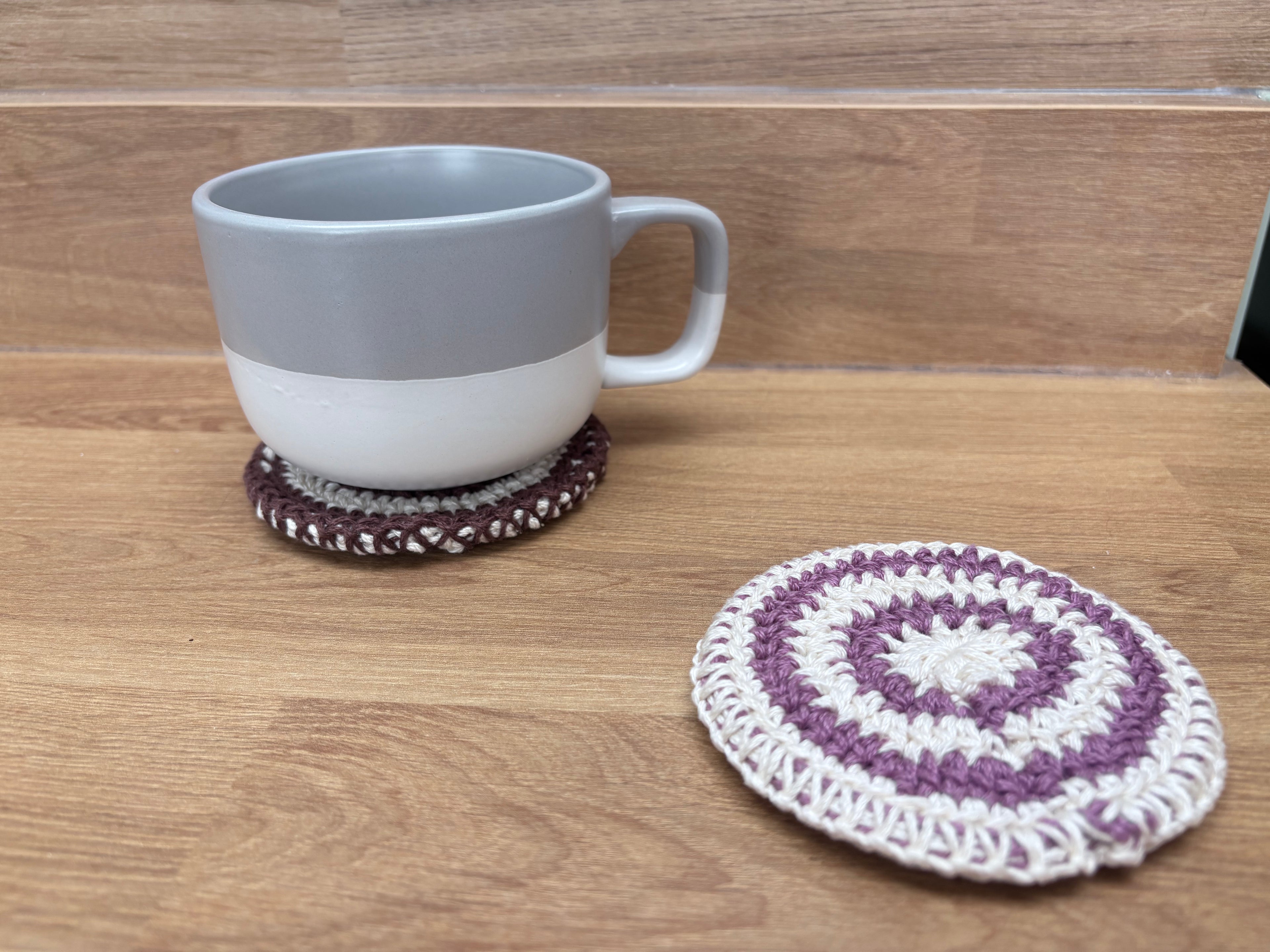 A gray and white ceramic mug sits on a round braided coaster, with another round purple and white crocheted coaster placed beside it on a wooden tabletop.