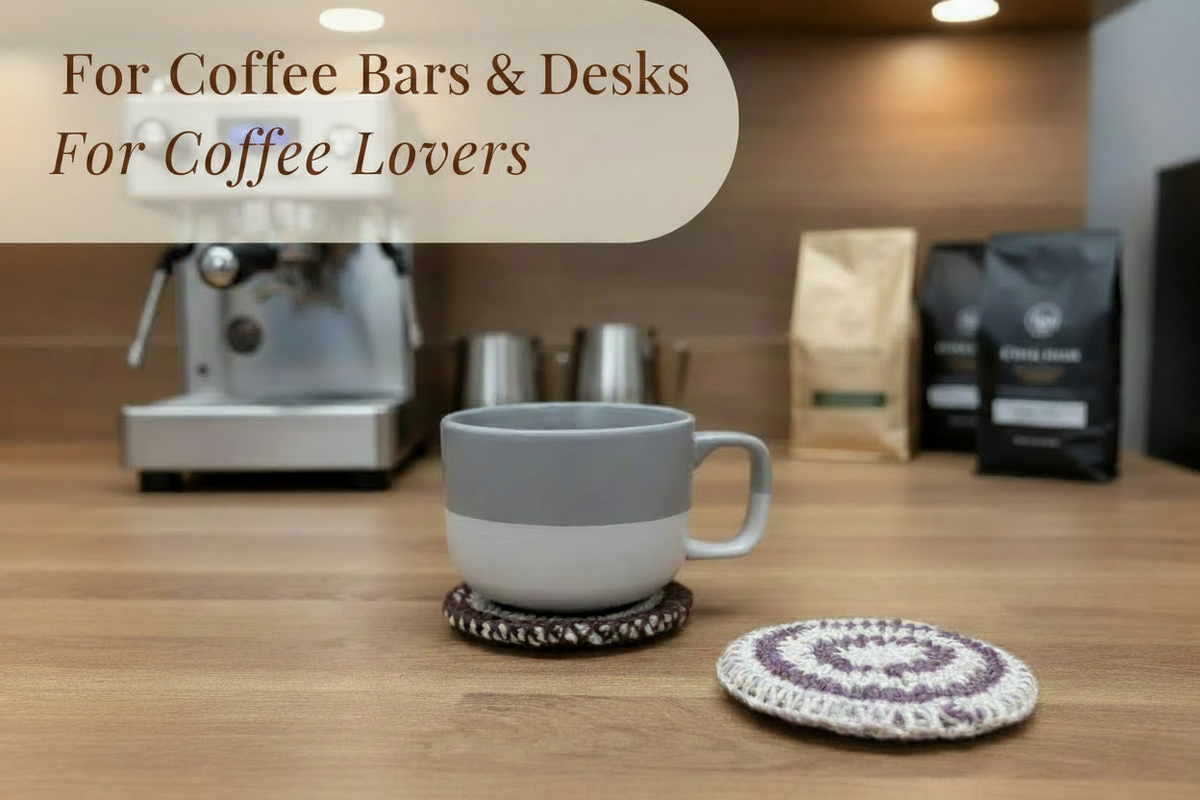A gray and white ceramic coffee mug sits on a wooden desk in a cafe setting, with a crocheted coaster in front of it. In the background, an espresso machine is visible, along with coffee bags.