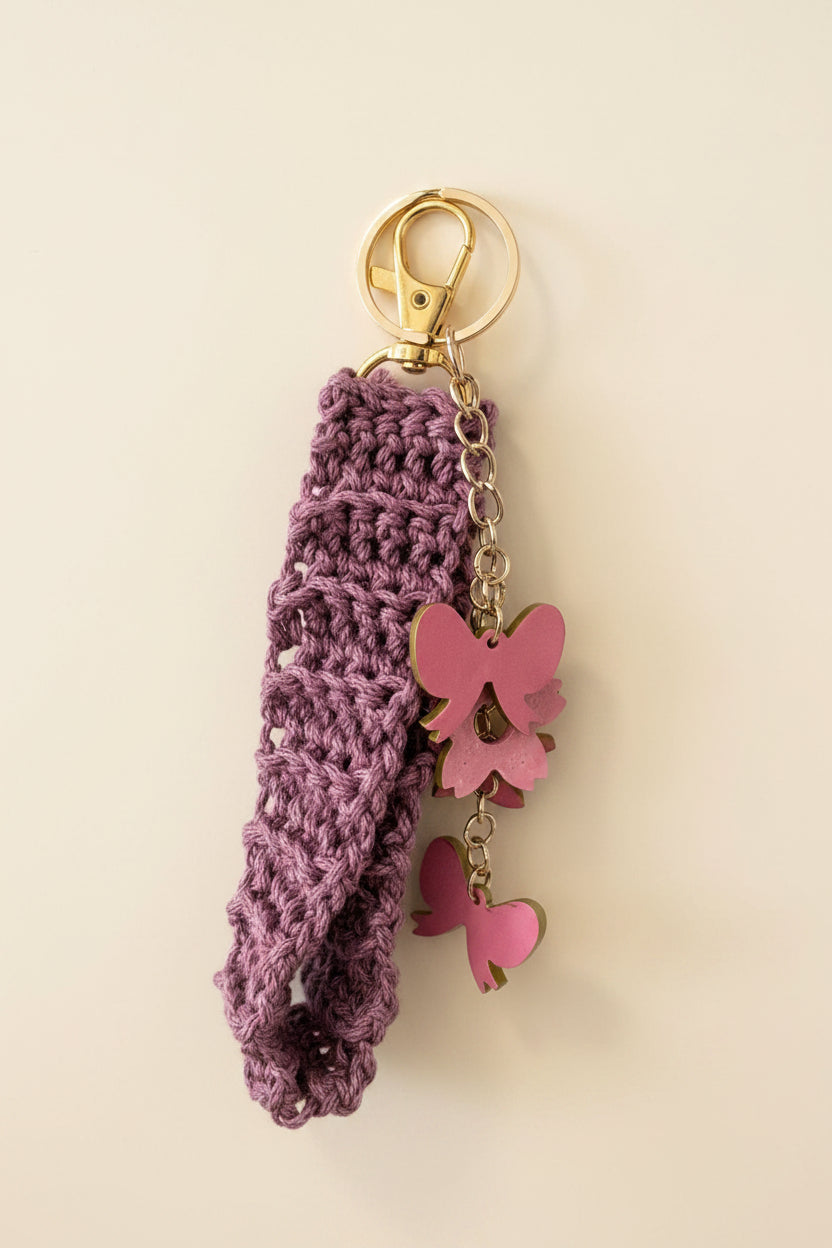 A purple crocheted wristlet strap attached to a gold-tone key ring and clasp, with pink butterfly and heart charms, hanging against a light gray wall.