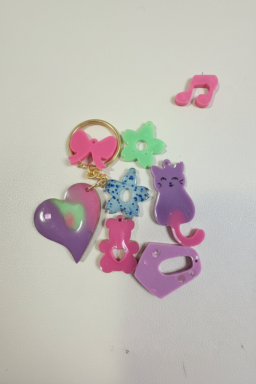 A colorful acrylic keychain featuring multiple cute shapes including a purple cat, pink bow, green flower, blue patterned flower, pink bear, and purple tag, displayed on a white desk.