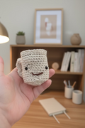 A hand holding a small crocheted mug with a cute face, featuring black bead eyes and a stitched smile. The background shows a blurred kitchen setting with a wooden counter and a painting on the wall.